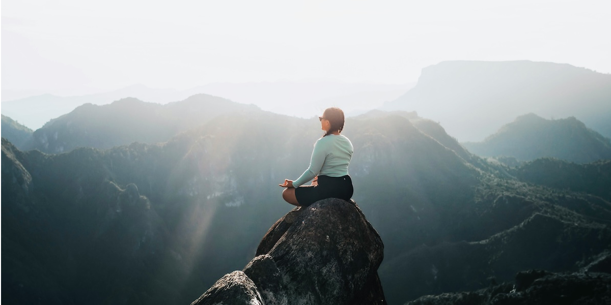 Person meditating on a rocky mountain peak overlooking misty ridges illuminated by sunlight.