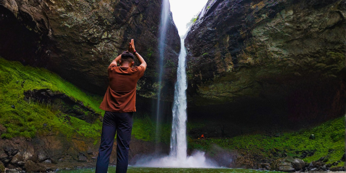 Man standing before a waterfall with hands raised in a prayer gesture, surrounded by moss-covered cliffs.